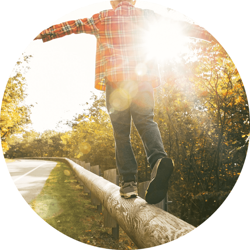 Child balancing on a wooden rail, arms outstretched, with sunlight shining through trees along a quiet road.