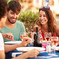 People dining outdoors, eating and laughing, seated at a table with wine glasses and colorful drinks.